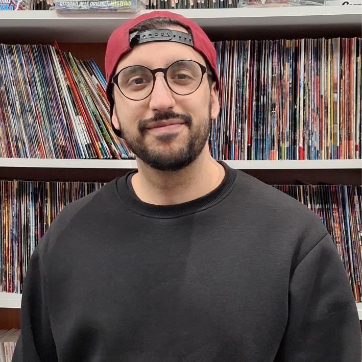 Man wearing a black sweatshirt and red cap in front of a bookshelf filled with books.