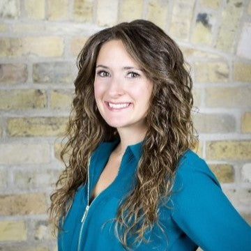 Woman with curly hair wearing a teal shirt against a brick wall.
