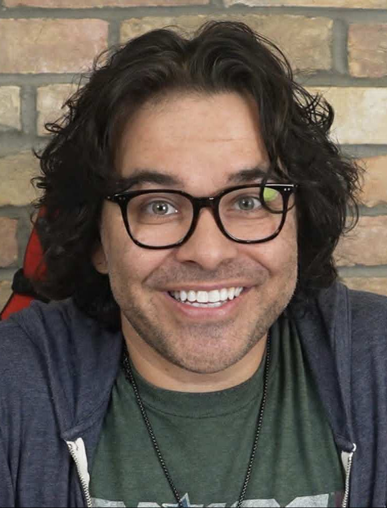 Man with glasses and a hoodie smiling in front of a brick wall